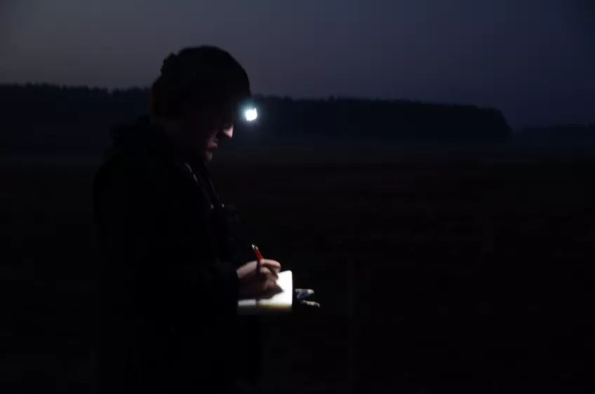 En person med pannlampa står i mörker och skriver i en anteckningsbok. Foto.